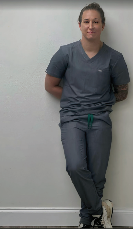 A woman in scrubs leans against a wall with a visible tattoo on left arm